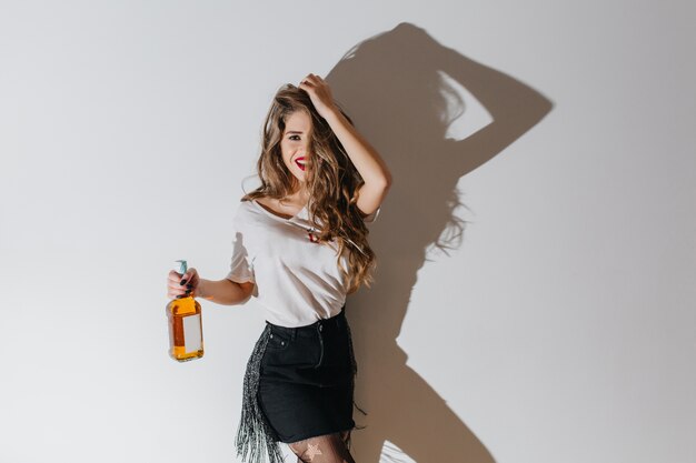 Slim femme qui rit avec une coiffure longue debout en studio avec une bouteille de champagne