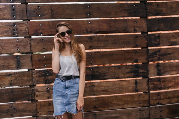 Slim jeune femme posant sur un mur en bois et touchant ses lunettes de soleil. Portrait en plein air d'une fille caucasienne extatique porte une jupe en jean.