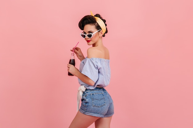Slim pin-up en short en jean à la recherche par-dessus l'épaule. Photo de Studio de la belle jeune femme avec une bouteille de soda debout sur fond rose.