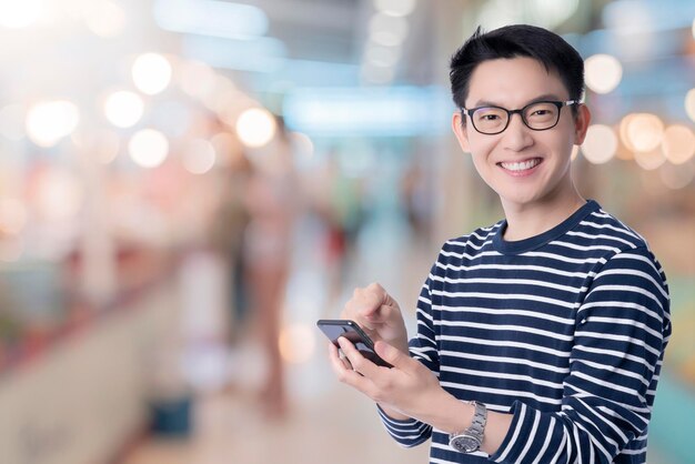 Smart casual bonheur asiatique mâle porter des lunettes sourire et main joyeuse utiliser smartphone prêt à appuyer sur le bouton d'achat avec centre commercial abstrait flou fond technologie communication idées concept