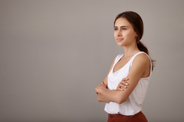 Smart mignonne belle jeune femme avec charmant sourire heureux posant dans une posture confiante avec les bras croisés sur sa poitrine, regardant devant elle vers un avenir meilleur et brillant, ayant une expression de rêve