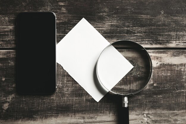 Smartphone mobile, loupe et feuille de papier blanc isolé sur table en bois de ferme noire Concept de jeu de détective mystérieux.