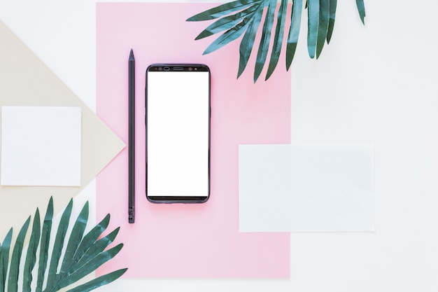 Photo gratuite smartphone près de papiers et de palmiers sur un bureau blanc