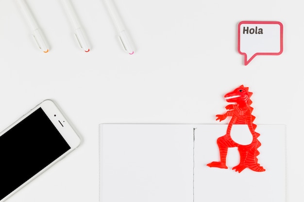 Smartphone près de stylo-feutre, papier, dinosaure jouet et cadre avec inscription Hola