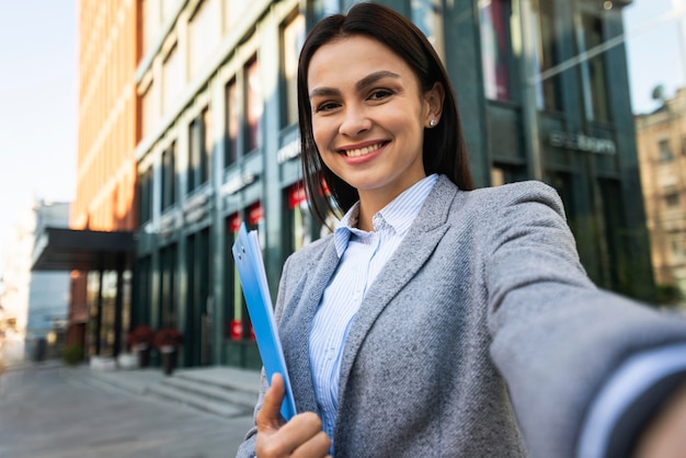 Smiley businesswoman avec presse-papiers en prenant un selfie à l'extérieur