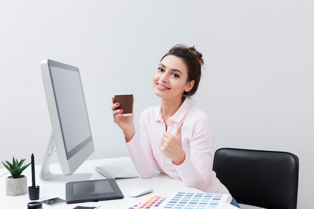 Smiley femme au bureau tenant une tasse de café et donner les pouces vers le haut