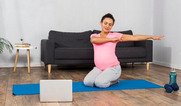 Smiley femme enceinte à la maison exercice sur tapis avec ordinateur portable