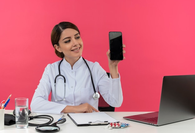 Smilin jeune femme médecin portant une robe médicale avec stéthoscope assis au bureau de travail sur ordinateur avec des outils médicaux holding phone with copy space