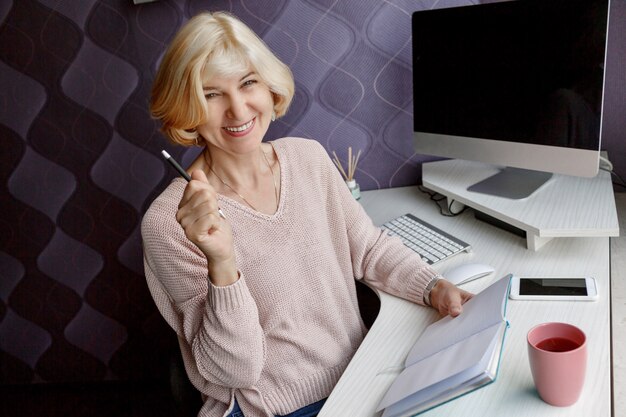 Smiling blonde mature woman écrit dans son agenda tout en travaillant par ordinateur à la maison