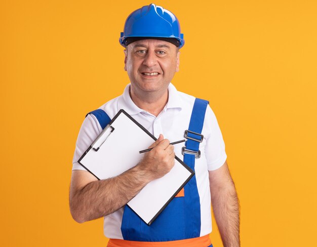 Photo gratuite smiling caucasian adult builder homme en uniforme tient un crayon et un presse-papiers sur orange