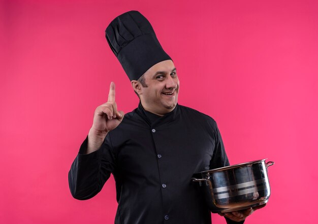 Smiling male d'âge moyen cuisinier en uniforme de chef holding casserole pointe du doigt vers le haut avec copie espace