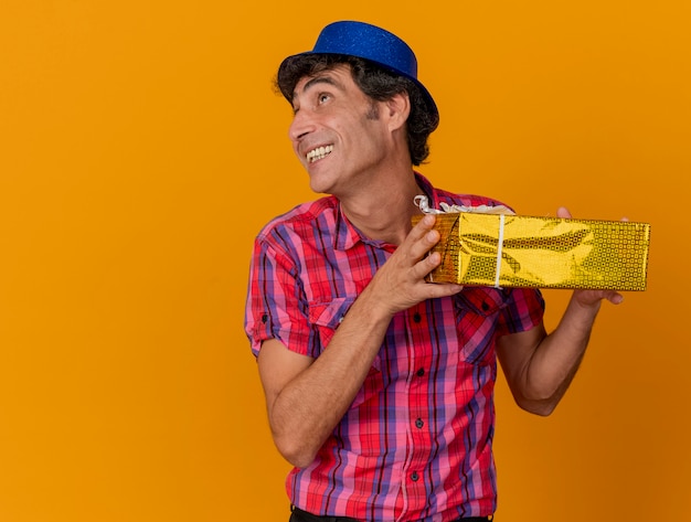 Smiling middle-age caucasian party man wearing party hat holding gift pack looking at side isolé sur fond orange avec copie espace