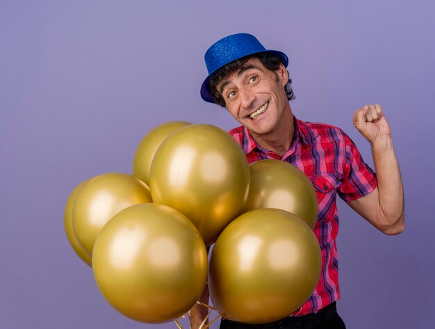 Smiling party man wearing party hat holding balloons looking at side faisant oui geste isolé sur mur violet
