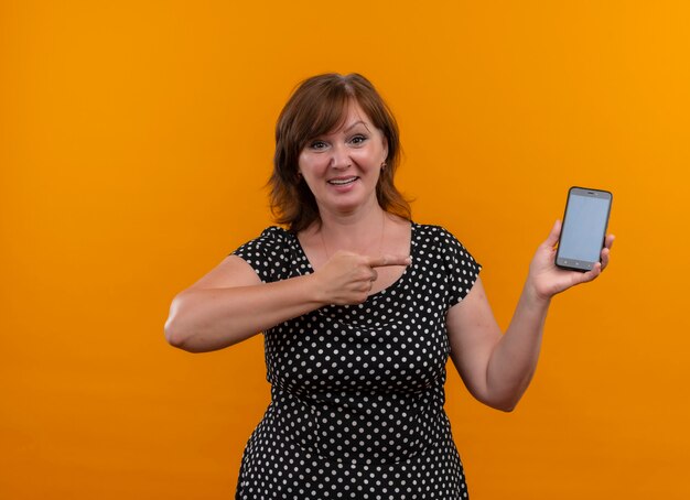 Smiling woman holding mobile phone et pointant avec le doigt dessus sur un mur orange isolé avec copie espace