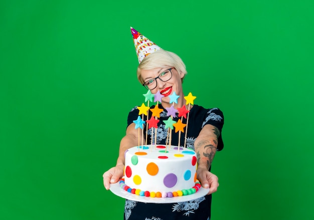 Smiling young blonde party girl portant des lunettes et une casquette d'anniversaire qui s'étend du gâteau d'anniversaire avec des étoiles regardant la caméra isolée sur fond vert avec espace de copie