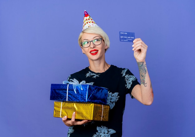 Smiling Young Blonde Party Girl Portant Des Lunettes Et Une Casquette D'anniversaire Tenant Des Coffrets Cadeaux Et Une Carte De Crédit En Regardant La Carte Isolée Sur Fond Violet Avec Copie Espace