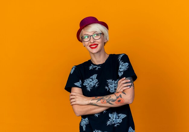 Smiling young blonde party girl wearing party hat et lunettes debout avec une posture fermée regardant côté isolé sur fond orange avec copie espace