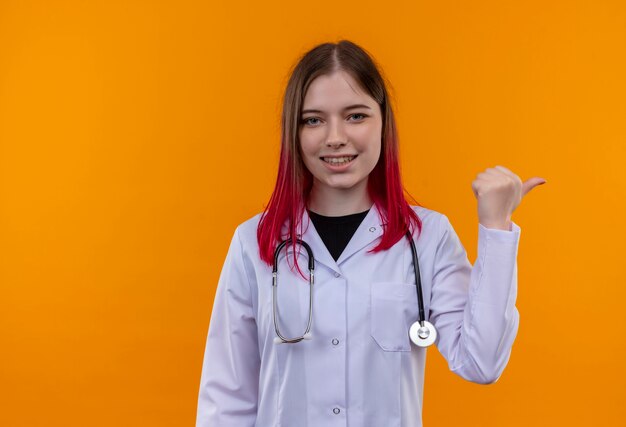 Smiling young doctor girl wearing stéthoscope robe médicale pointe à côté sur fond orange isolé avec copie espace