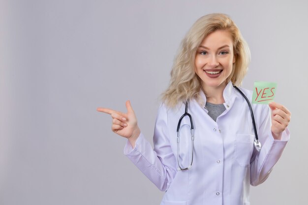 Smiling young doctor wearing stethoscope in medical gown holding paper oui marque et pointe vers le côté sur le mur blanc