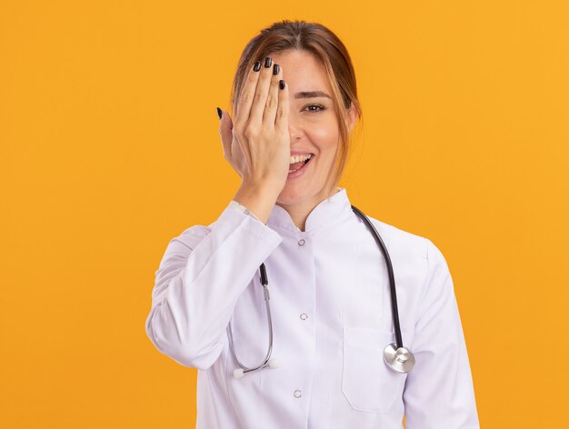 Smiling young female doctor wearing medical robe avec stéthoscope couvert oeil avec main isolé sur mur jaune