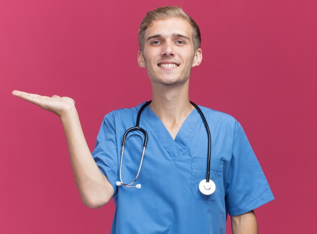Smiling young male doctor wearing doctor uniform avec stéthoscope faisant semblant de tenir quelque chose d'isolé sur un mur rose avec copie espace