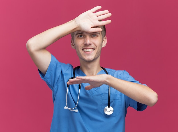 Smiling young male doctor wearing doctor uniform avec stéthoscope mettant la main sur le front isolé sur mur rose