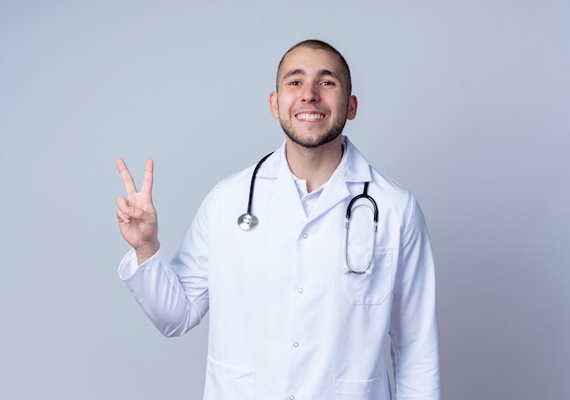 Smiling young male doctor wearing medical robe et stéthoscope autour de son cou faisant signe de paix isolé sur mur blanc