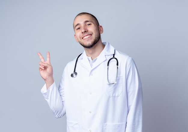 Smiling young male doctor wearing medical robe et stéthoscope faisant signe de paix autour de son cou isolé sur mur blanc