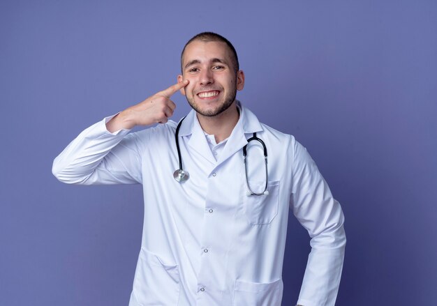 Smiling young male doctor wearing medical robe et stéthoscope mettant le doigt sur la joue isolé sur le mur violet