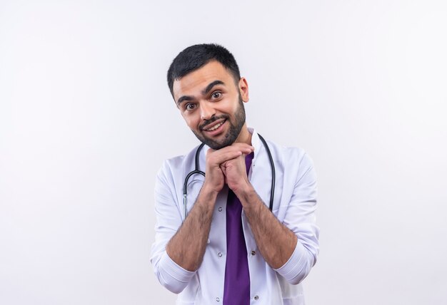 Smiling young male doctor wearing stethoscope medical gown mis ses mains sur le menton sur un mur blanc isolé