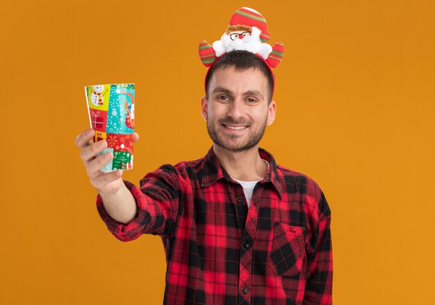 Smiling young man wearing santa claus headband étendant la tasse de Noël en plastique vers la caméra en regardant la caméra isolée sur fond orange avec copie espace