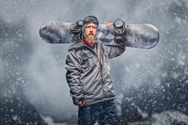 Snowboarder rousse brutal avec une barbe pleine dans un chapeau d'hiver et des lunettes de protection vêtu d'un manteau de snowboard posant avec un snowboard sur fond de montagnes, regardant une caméra.