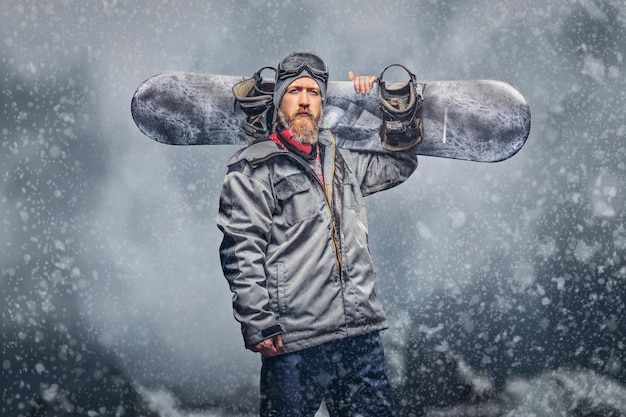 Snowboarder rousse brutal avec une barbe pleine dans un chapeau d'hiver et des lunettes de protection vêtu d'un manteau de snowboard posant avec un snowboard sur fond de montagnes, regardant une caméra.