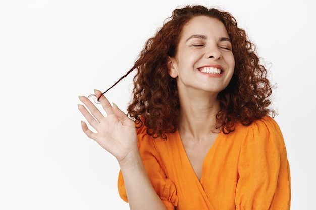 Soins de la peau de beauté. Portrait de jeune femme naturelle aux cheveux bouclés rouges, riant heureux, dents de sourire blanches, jouant avec les cheveux, debout insouciant sur fond blanc