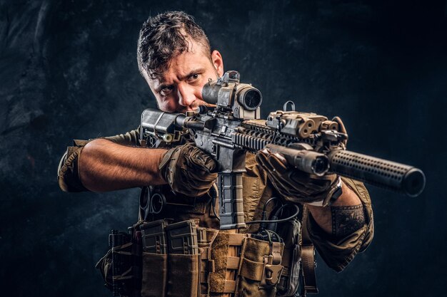 Soldat des forces spéciales portant une armure corporelle tenant un fusil d'assaut et visant l'ennemi. Photo de studio contre un mur texturé sombre