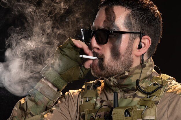 Soldat masculin en camouflage fumant une cigarette sur un mur noir