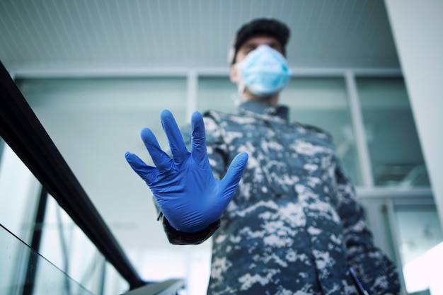 Soldat en uniforme de camouflage portant des gants de protection et un masque montrant un panneau d'arrêt à l'entrée de l'hôpital