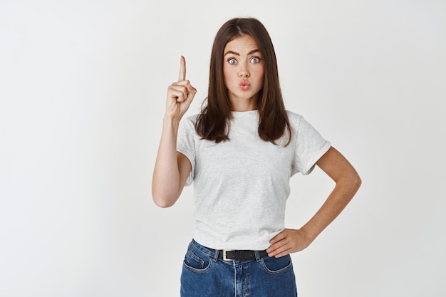 Solution. Jeune femme à la mode ayant une idée, pointant le doigt vers le haut et les lèvres plissées excitées, suggérant un plan, debout sur un mur blanc.