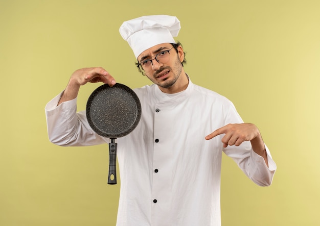 Sonfused jeune homme cuisinier portant l'uniforme de chef et des verres tenant et pointe vers une poêle sur vert