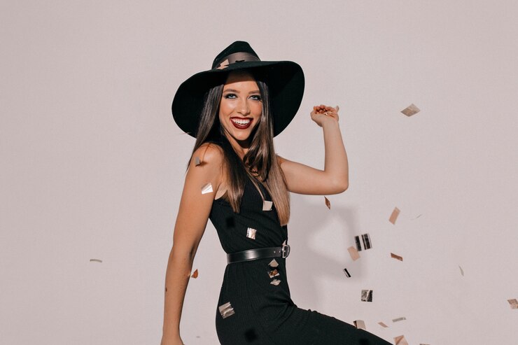 Sortie de femme brune riante active au chapeau noir et élégante robe noire avec des lèvres sombres en attente de fête