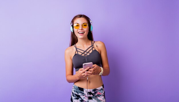 Sortie de femme sportive avec un sourire sincère posant dans des vêtements actifs élégants en studio. Utiliser un smartphone et écouter de la musique.