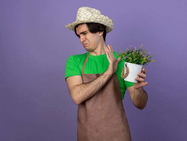 Soucieux jeune jardinier mâle en uniforme portant chapeau de jardinage tenant une fleur en pot de fleurs