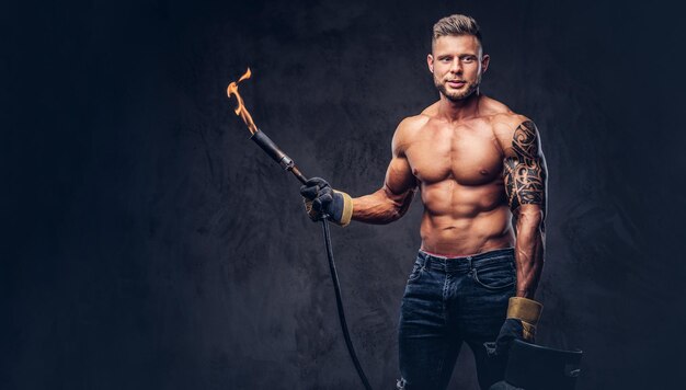 Soudeur mâle tatoué brutal avec une coupe de cheveux et une barbe élégantes, avec un corps musclé, vêtu uniquement de jeans, tient un réservoir de propane et un brûleur brûlant, debout dans un studio, regardant une caméra. Isolé sur