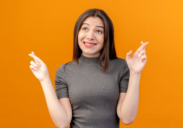 Souhaitant jeune jolie femme croisant les doigts et levant isolé sur fond orange