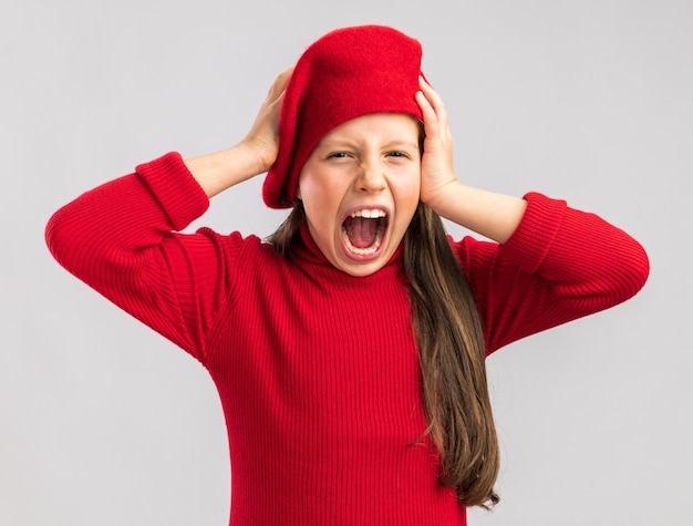 A souligné une petite fille blonde portant un béret rouge en gardant les mains sur la tête en regardant la caméra et en criant isolé sur un mur blanc