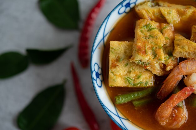 Soupe aigre-douce avec cha-om, œuf et crevettes dans un bol blanc, avec chili et feuilles de lime kaffir sur une surface blanche.