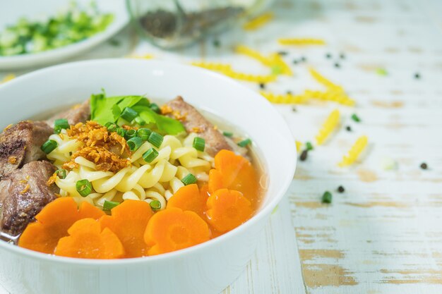Soupe de macaronis au porc et carottes sur une table en bois blanche