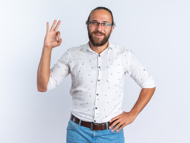 Souriant adulte bel homme portant des lunettes gardant la main sur la taille en regardant la caméra faisant signe ok isolé sur mur blanc