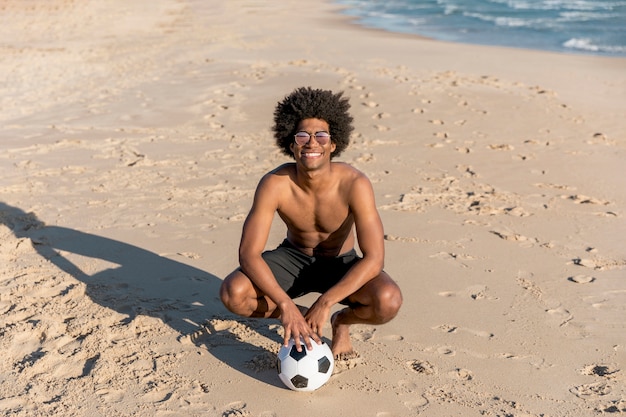 Photo gratuite souriant afro-américain assis avec ballon sur la plage d'été