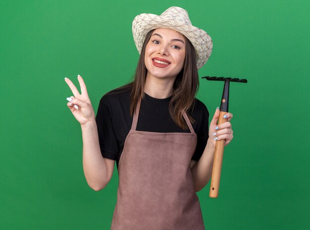 Souriant assez caucasien femme jardinier portant chapeau de jardinage tenant râteau et signe de victoire gesticulant isolé sur mur vert avec espace copie